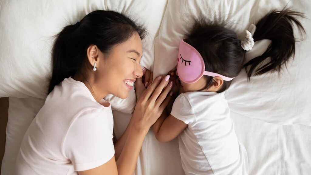 Top above view smiling caring asian ethnic mommy watching little preschool biracial baby girl enjoying good night time on soft pillow, wearing sleeping mask, relaxing together on comfortable bed.